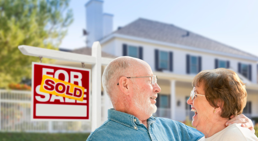 Happy Affectionate Senior Couple Hugging in Front of Sold Real Estate Sign and House.